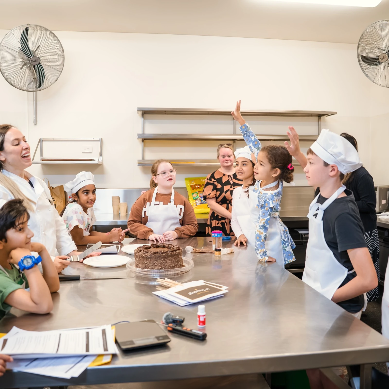 A chef and a group of children in aprons and chef hats around a kitchen bench, some with their hands raised