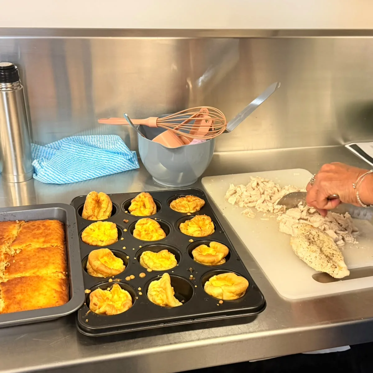 A kitchen bench with a tray of baked cornbread, a tray of Yorkshire puddings and a participant slicing cooked chicken on a board